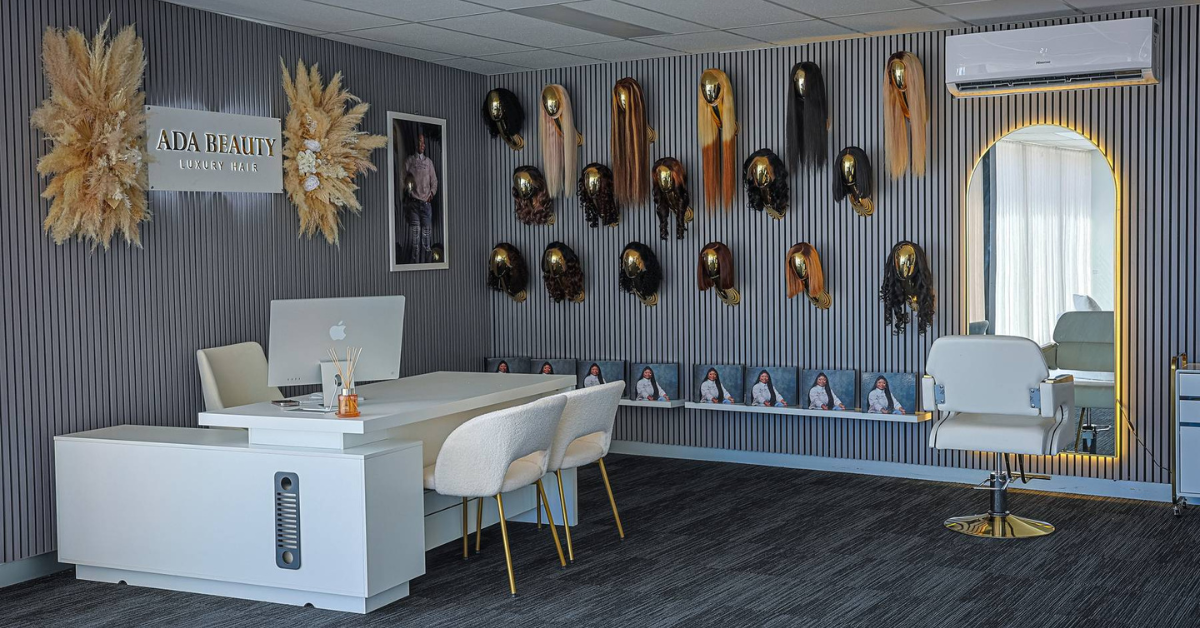 Modern hair salon interior with wigs on a striped wall, white desk and chairs, and a styling chair at the workstation.