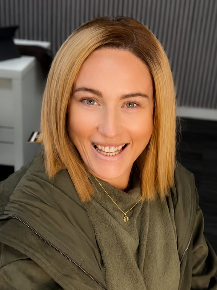 Close-up portrait of a smiling woman with shoulder-length blonde hair wearing a green jacket in an indoor office setting